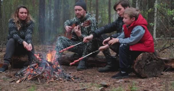 父亲正在弹吉他 而他的孩子和妻子在煎肉 一家人围坐在篝火边 孩子们和他们的父母都在森林里休息 家庭在森林的火堆上煎香肠 — 图库视频影像