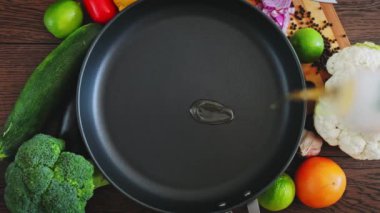 Aşçı tavaya ayçiçeği yağı döker, üst manzara. Mutfak tavası kahverengi tahtaların üzerinde duruyor, etrafında sebzeler ve yağlar kızartılması için içine dökülüyor. Kızartma tavasına dökülen yağın yavaş hareketi. 