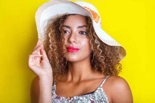 Beauty portrait of young african american girl with afro hairstyle in summer hat. Girl posing on yellow background, looking at camera. — Stock Photo, Image