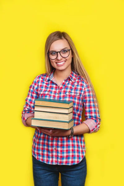 Smiling young stylish student is standing with books on yellow background in glasses and casual bright outfit — Stock Photo, Image