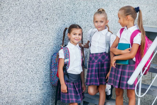 Pupils of primary school. Girls with backpacks near building outdoors. Beginning of lessons. First day of fall.
