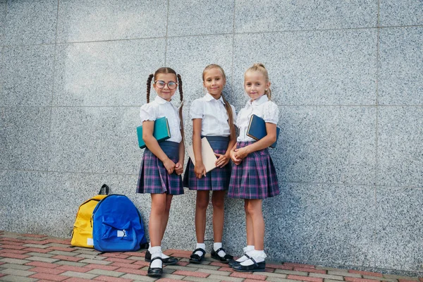 Pupils of primary school. Girls with backpacks and books near building outdoors. Beginning of lessons. First day of fall.