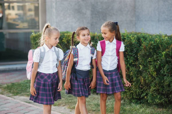 Pupils of primary school. Girls with backpacks near building outdoors. Beginning of lessons. First day of fall.