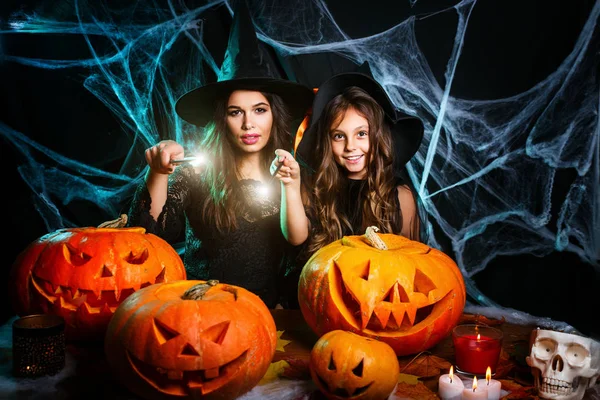 Vrolijke Halloween. Mooie Kaukasische moeder en haar dochter in heks kostuums genieten met behulp van magie met toverstaf aan halloween pompoen jar over spinnenweb op zwarte studio achtergrond. — Stockfoto