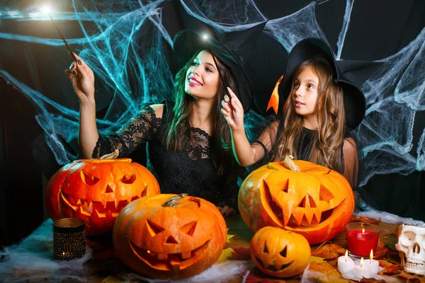 Vrolijke Halloween. Mooie Kaukasische moeder en haar dochter in heks kostuums genieten met behulp van magie met toverstaf aan halloween pompoen jar over spinnenweb op zwarte studio achtergrond. — Stockfoto