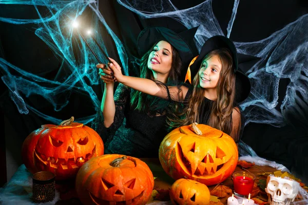 Vrolijke Halloween. Mooie Kaukasische moeder en haar dochter in heks kostuums genieten met behulp van magie met toverstaf aan halloween pompoen jar over spinnenweb op zwarte studio achtergrond. — Stockfoto