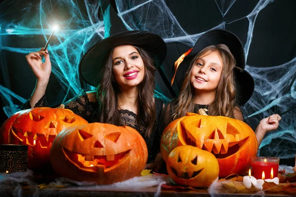 Vrolijke Halloween. Mooie Kaukasische moeder en haar dochter in heks kostuums genieten met behulp van magie met toverstaf aan halloween pompoen jar over spinnenweb op zwarte studio achtergrond. — Stockfoto