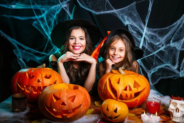 Fröhliches Halloween. niedliches kleines Mädchen und ihre schöne Mutter in Hexenhüten schauen in die Kamera und lächeln, während sie auf einem für Halloween dekorierten Holzhintergrund sitzen — Stockfoto