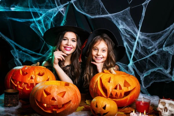 Fröhliches Halloween. niedliches kleines Mädchen und ihre schöne Mutter in Hexenhüten schauen in die Kamera und lächeln, während sie auf einem für Halloween dekorierten Holzhintergrund sitzen — Stockfoto