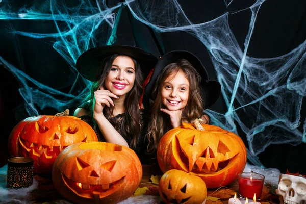 Buon Halloween. Carino bambina e la sua bella mamma in cappelli strega stanno guardando la fotocamera e sorridente mentre seduto su uno sfondo di legno decorato per Halloween — Foto Stock