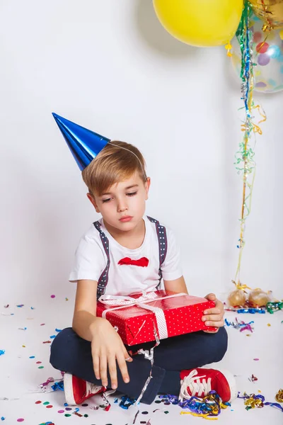 Cute sad little boy in birthday cap holding red present. Holidays concept.