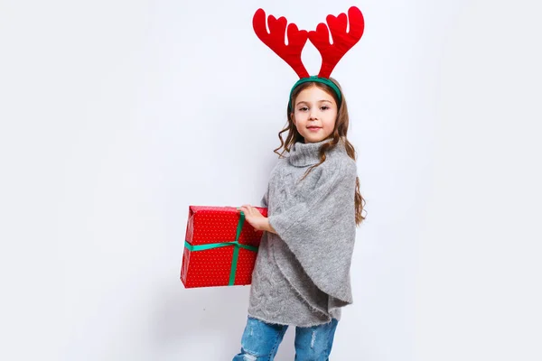 Счастливая маленькая улыбчивая девочка с подарочной коробкой, смотрящая в камеру. Рождественская идея. Улыбающаяся смешная девушка в оленьих рогах в студии . — стоковое фото