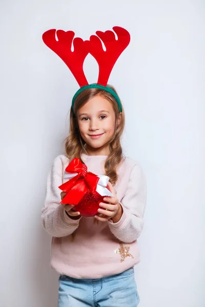 Feliz niña sonriente con bola de árbol de Navidad rojo. Concepto de Navidad. Sonriente chica divertida en cuernos de ciervo sobre fondo blanco —  Fotos de Stock