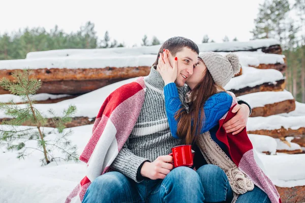 Jovem casal amoroso vestido com suéter azul sentado com xícaras de café vermelho na lenha na floresta de inverno — Fotografia de Stock