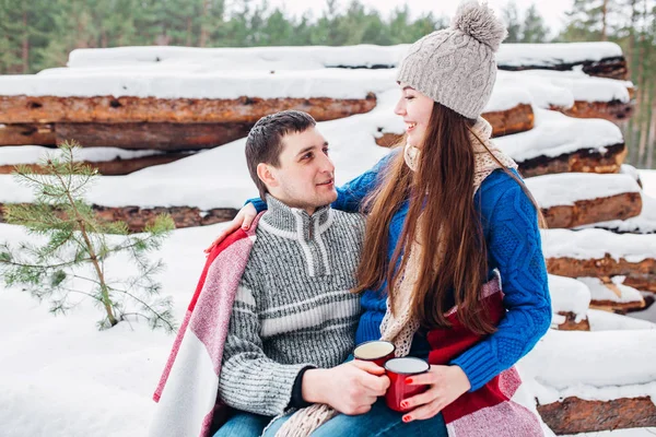 Jovem casal amoroso vestido com suéter azul sentado com xícaras de café vermelho na lenha na floresta de inverno — Fotografia de Stock