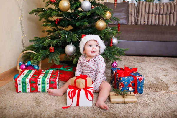 Feliz linda niña en Santa Claus sombrero celebración de regalos de Navidad en el árbol de Navidad en casa —  Fotos de Stock