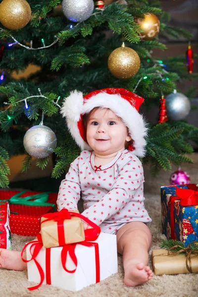 Feliz linda niña en Santa Claus sombrero celebración de regalos de Navidad en el árbol de Navidad en casa —  Fotos de Stock