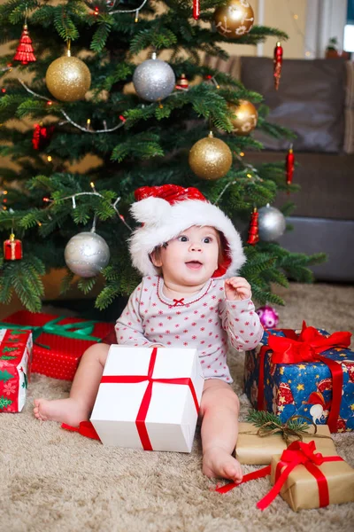 Feliz linda niña en Santa Claus sombrero celebración de regalos de Navidad en el árbol de Navidad en casa —  Fotos de Stock