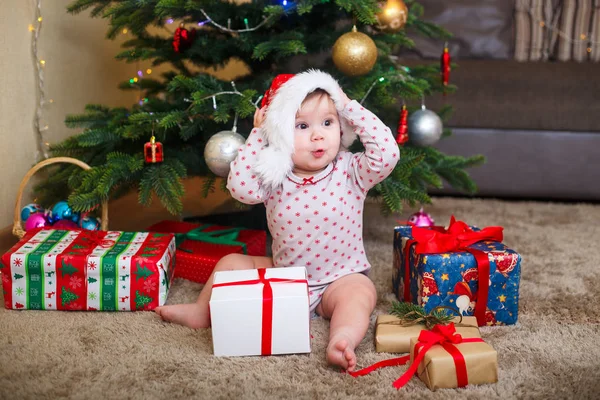 Feliz linda niña en Santa Claus sombrero celebración de regalos de Navidad en el árbol de Navidad en casa —  Fotos de Stock