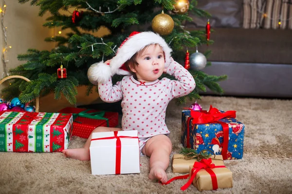Feliz linda niña en Santa Claus sombrero celebración de regalos de Navidad en el árbol de Navidad en casa —  Fotos de Stock