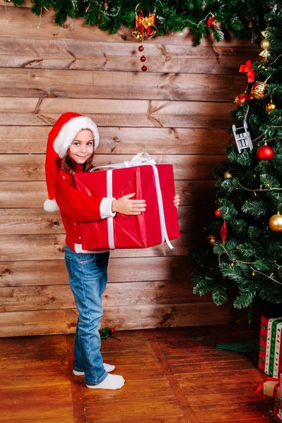 Feliz Navidad y Felices Fiestas. Linda niña con una gran caja de regalo roja cerca del árbol interior —  Fotos de Stock