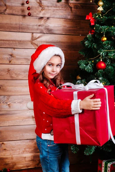 Feliz Navidad y Felices Fiestas. Linda niña con una gran caja de regalo roja cerca del árbol interior —  Fotos de Stock