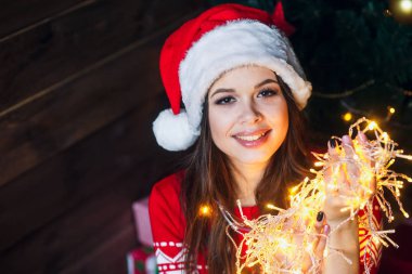 Güzel genç kadın gülümseyerek ve çelenk böke ışıklı Noel arka plan, ışıkların konuşurken Noel Baba şapkası