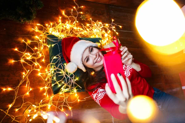 Retrato de cerca de la mujer en el sombrero de Santa envuelta en luces de Navidad tomando selfie y acostado en el suelo de madera en casa — Foto de Stock