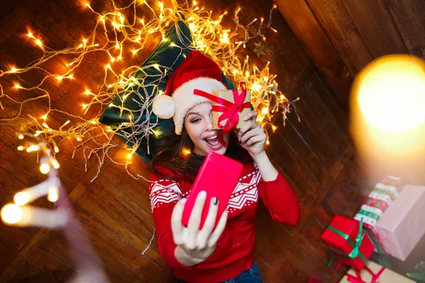 Retrato de cerca de la mujer en el sombrero de Santa envuelta en luces de Navidad tomando selfie y acostado en el suelo de madera en casa — Foto de Stock