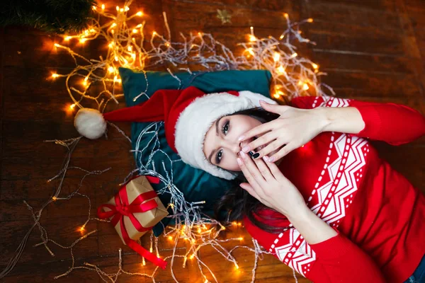 Primer plano retrato de la mujer en el sombrero de Santa envuelto en luces de Navidad divertirse y acostarse en el suelo — Foto de Stock