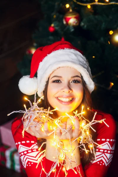 Belle jeune femme dans Santa chapeau souriant et parlant guirlandes de lumières à fond de Noël avec des lumières boke — Photo