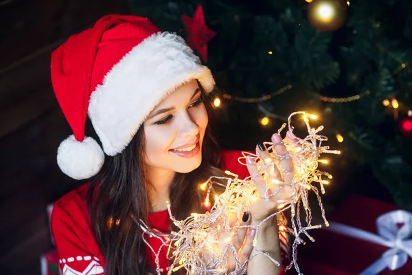 Hermosa joven en Santa sombrero sonriendo y hablando guirnaldas de luces en el fondo de Navidad con luces boke — Foto de Stock