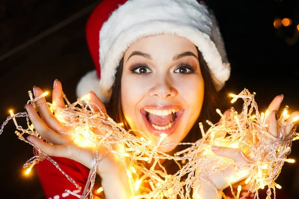 Hermosa joven en Santa sombrero sonriendo y hablando guirnaldas de luces en el fondo de Navidad con luces boke — Foto de Stock