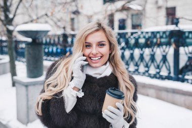 Gülümseyen genç kadın kürk paltolu kar yağışı Europe şehir merkezinde yürüyüş kahve ile çok etkileyici. Pozitifliği ifade, duyguları gerçek, kar yağışı, Noel tatili için bekleyen keyfini çıkarın