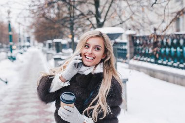 Gülümseyen genç kadın kürk paltolu kar yağışı Europe şehir merkezinde yürüyüş kahve ile çok etkileyici. Pozitifliği ifade, duyguları gerçek, kar yağışı, Noel tatili için bekleyen keyfini çıkarın