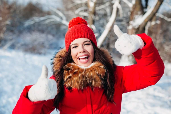 Mujer hermosa en ropa roja sonriendo a la cámara con los pulgares hacia arriba en el parque de invierno —  Fotos de Stock