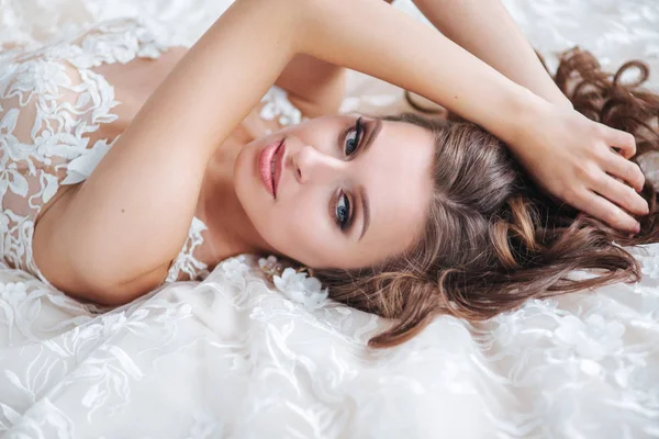 Vackra brud liggande på en vit säng på hotellet. Flickan i en vit klänning vilar i sovrummet — Stockfoto