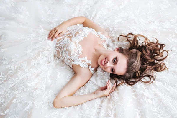 Vackra brud liggande på en vit säng på hotellet. Flickan i en vit klänning vilar i sovrummet — Stockfoto