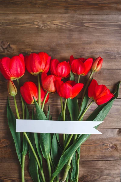 Lente bloemen. Boeket van rode tulpen met kaart voor tekst op bruin houten achtergrond. Mothers Day en Valentijnsdag concept. Plat leggen — Stockfoto