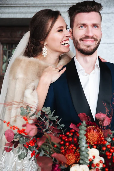 Feliz casal recém-casado posando e sorrindo em vestido de noiva e terno com flores — Fotografia de Stock