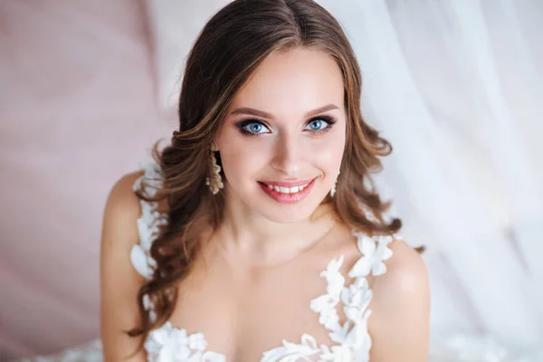 Hermosa novia con pelo rizado largo en vestido de novia elegante y diadema sonriendo en la habitación en la mañana de la boda — Foto de Stock