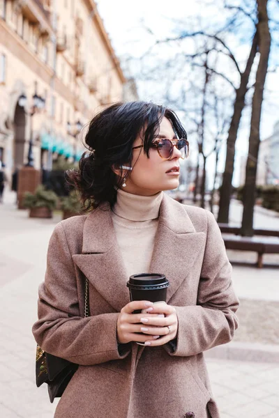 Beautiful elegant businesswoman in sunglasses and coat with airpods and coffee in city street — Stock Photo, Image
