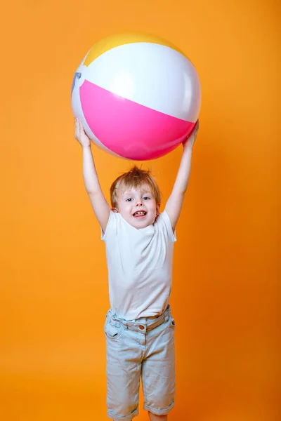 Cute Llttle boy in swimming shorts holding a beach ball on orange background