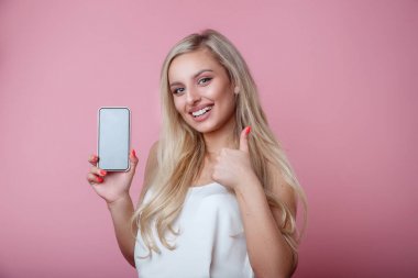 Güzel gülümseyen kadın başparmak gösteren ve akıllı telefon tutarak ve pembe arka plan üzerinde kameraya bakarak