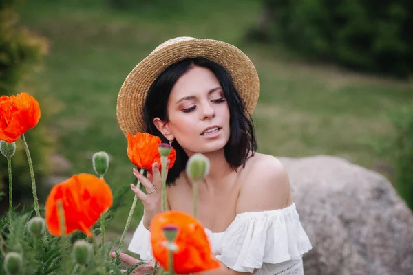 Romantyczna brunetka Kobieta w słomkowym kapeluszu z kwiatem w dłoni siedzi w niesamowitym polu maku. — Zdjęcie stockowe