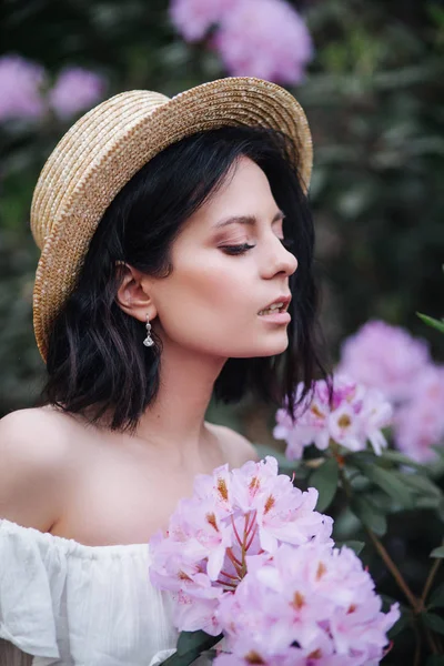 Retrato de cerca de una hermosa chica en un vestido vintage blanco y sombrero de paja de pie cerca de flores rosadas — Foto de Stock