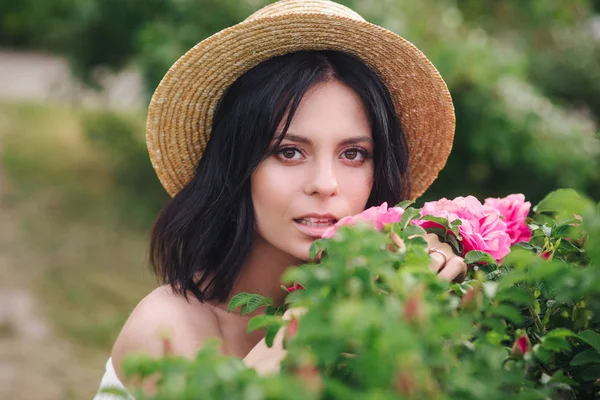 Romántico retrato de cerca o encantadora chica morena en sombrero de paja huele a flores en rosales — Foto de Stock