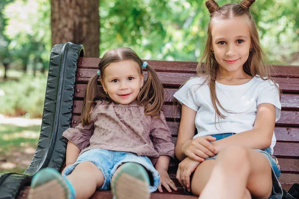 Dos niñas lindas sonriendo y sentadas en el banco de madera en el parque — Foto de Stock