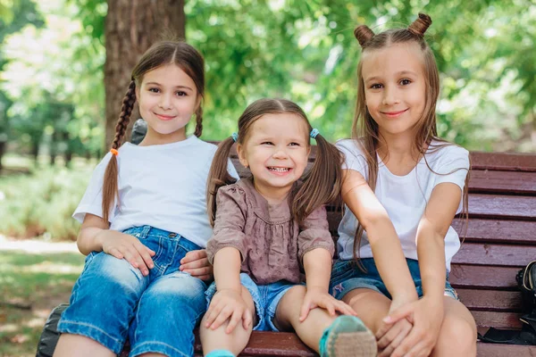 Tres niñas lindas sonriendo y sentadas en el banco de madera en el parque de verano — Foto de Stock