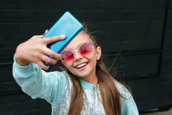 Güneş gözlüklü küçük güzel bir kız akıllı bir telefonla selfie çekiyor ve dışarıda gülümsüyor. — Stok fotoğraf